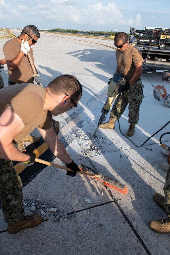 Three US Navy Sailors pictured repairing a runway. One operates a jackhammer while another sweeps with a push broom.