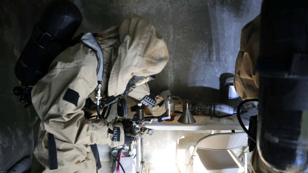 A U.S. Marine in chemical protective clothing checks places a purple chem light on a table with other equipment.