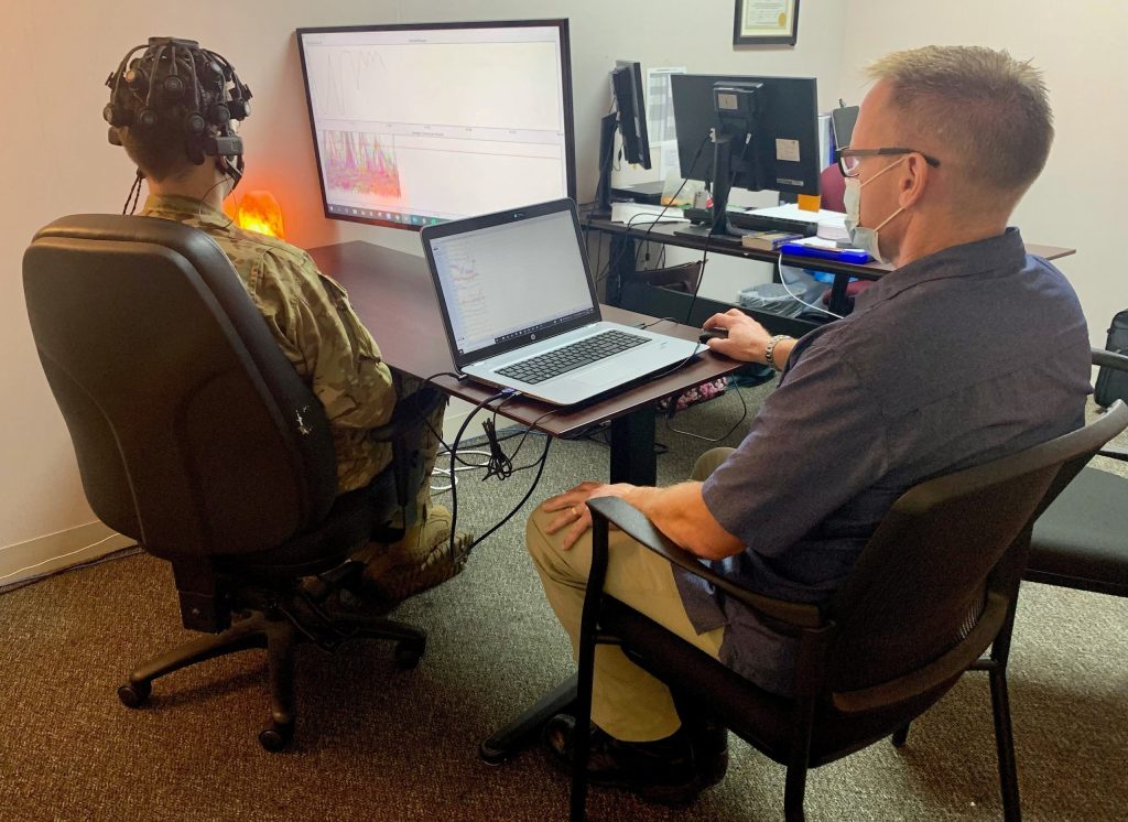 An Airman in uniform with sensors attached to his head sits at a computer screen. The test administrator, in civilian clothing, sits at a separate laptop.