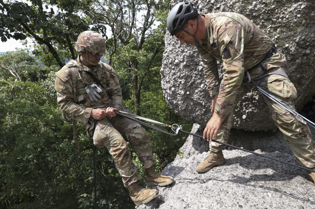 A Soldier prepares to rappel from a cliff. He is secured with a rope and leaning back getting ready to rappel down. Another Soldier looks on with his hand steadying the rope.