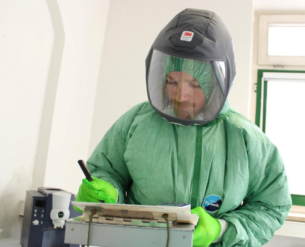 Department of Defense personnel in protective equipment writing on paper on a clipboard.