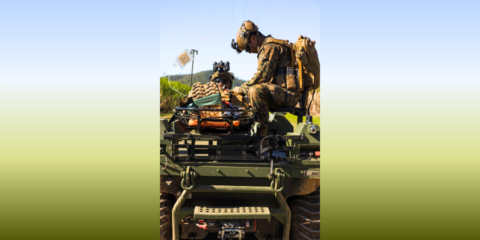 A Navy Hospital Corpsman sits beside a simulated patient on the back of a vehicle.