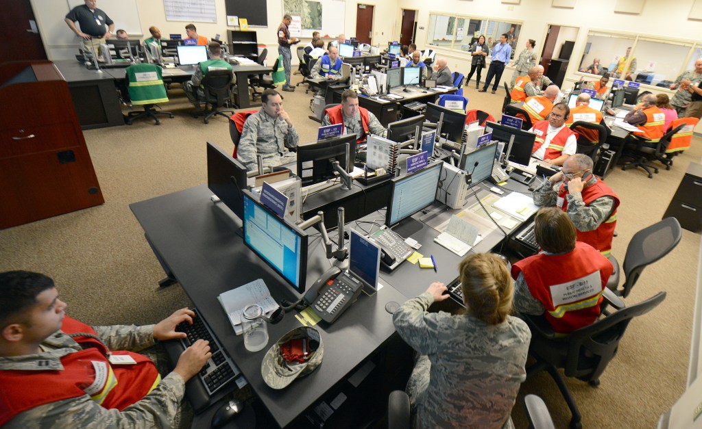 US Air Force Personnel staff an emergency operations center during a training exercise. Different groups are wearing different colored safety vests.