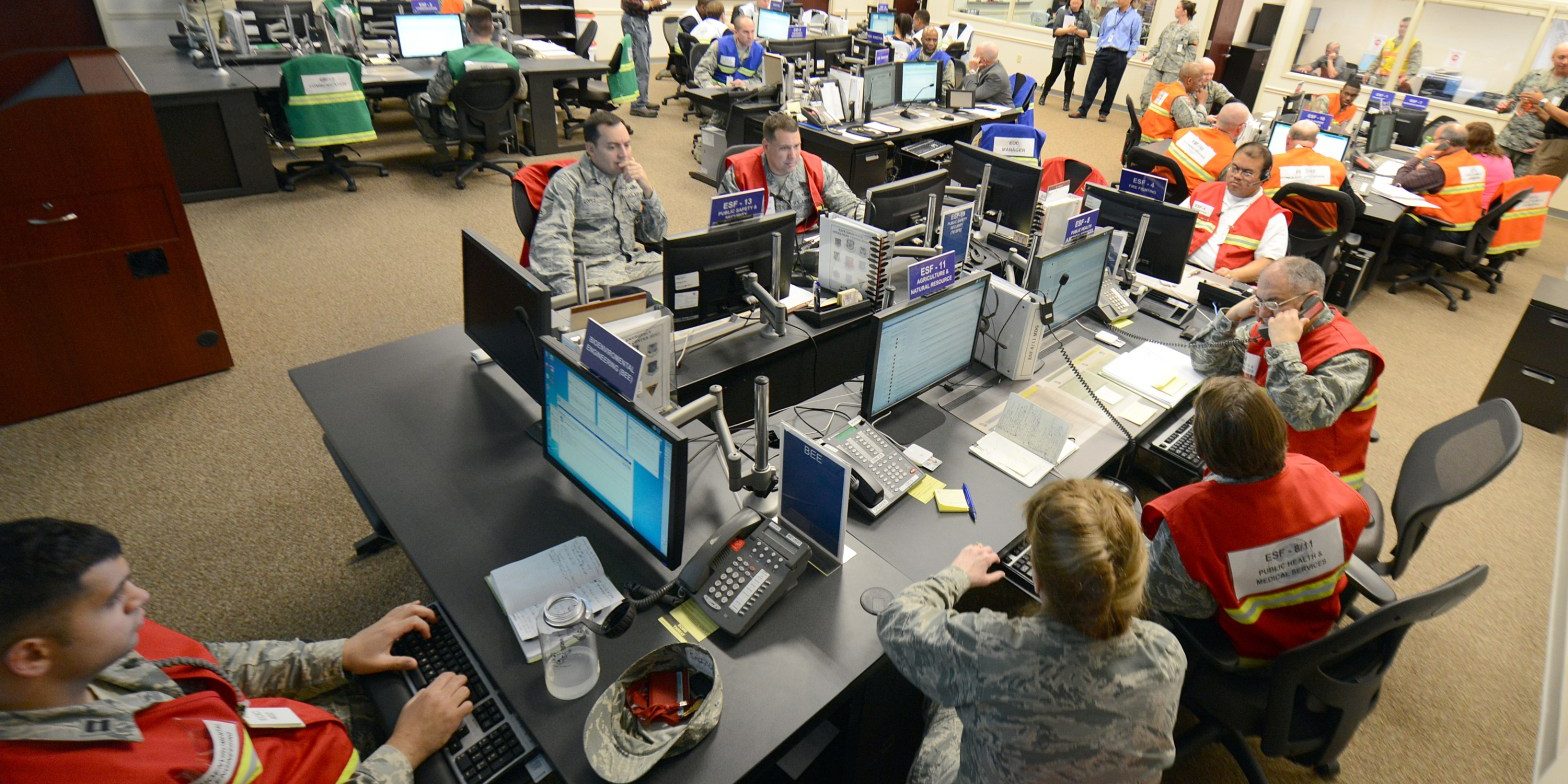 US Air Force Personnel staff an emergency operations center during a training exercise. Different groups are wearing different colored safety vests.