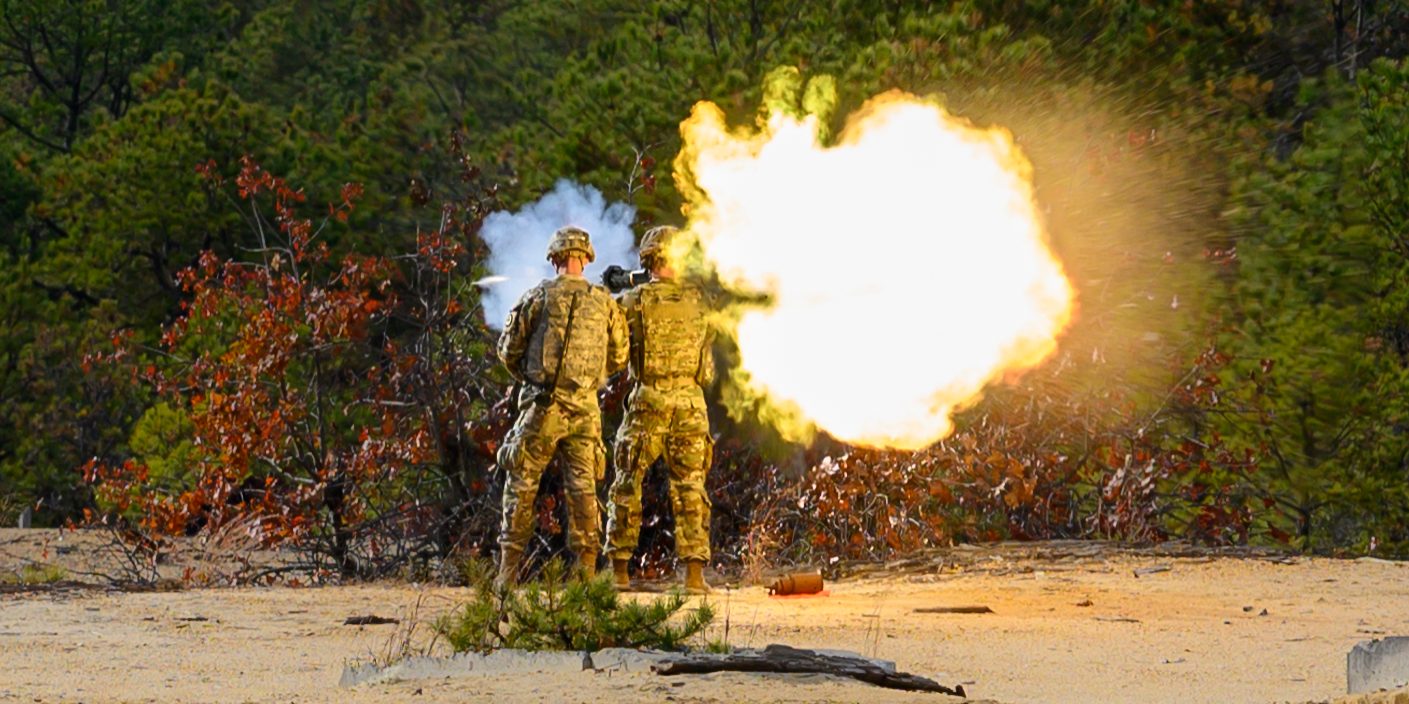 A soldier fires an AT-4 antitank rocket while another soldier observes. A large blast is shown behind the launcher and the projectile can be seen coming out of the front of the weapon.